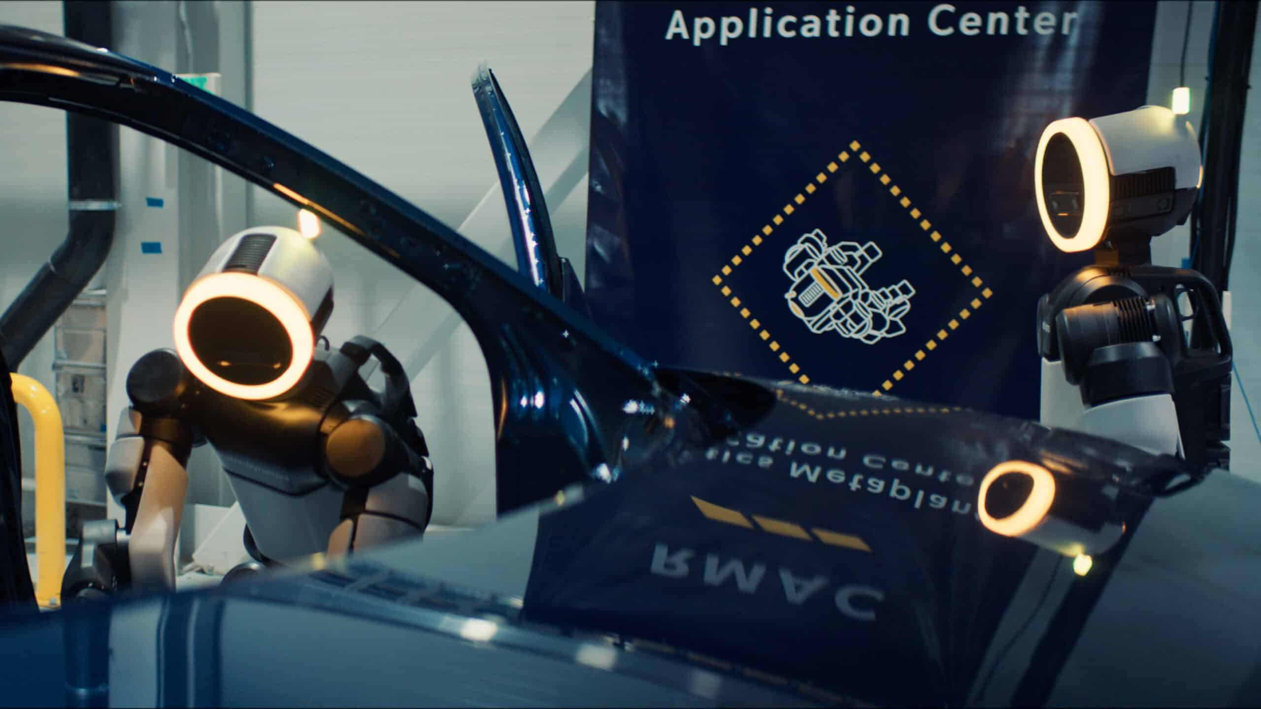 Two Atlas humanoid robots train around an automotive body panel inside the Robotics Metaplant Application Center.